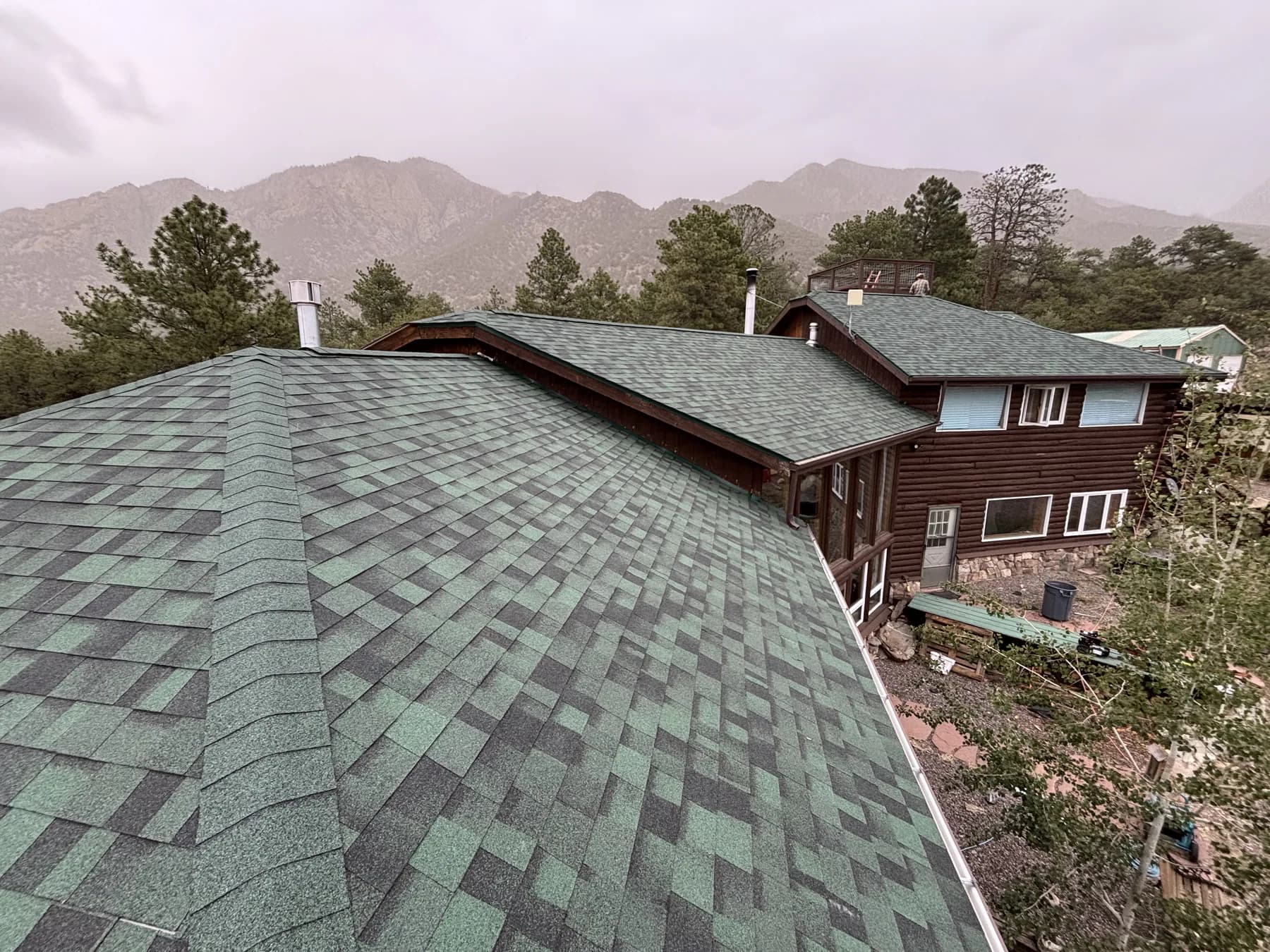 Green asphalt-shingle roof planes on a mountain home with peaks behind them.