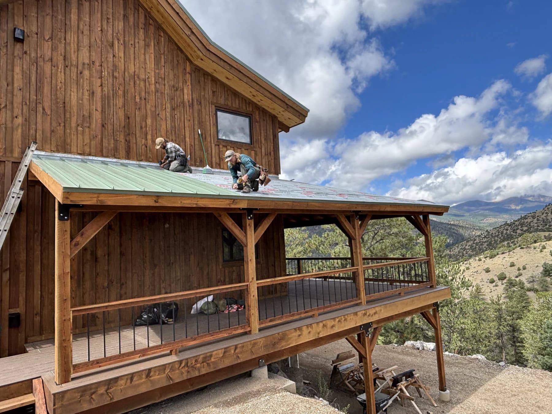 Two crew members working on a green metal porch roof beside a timber-sided home