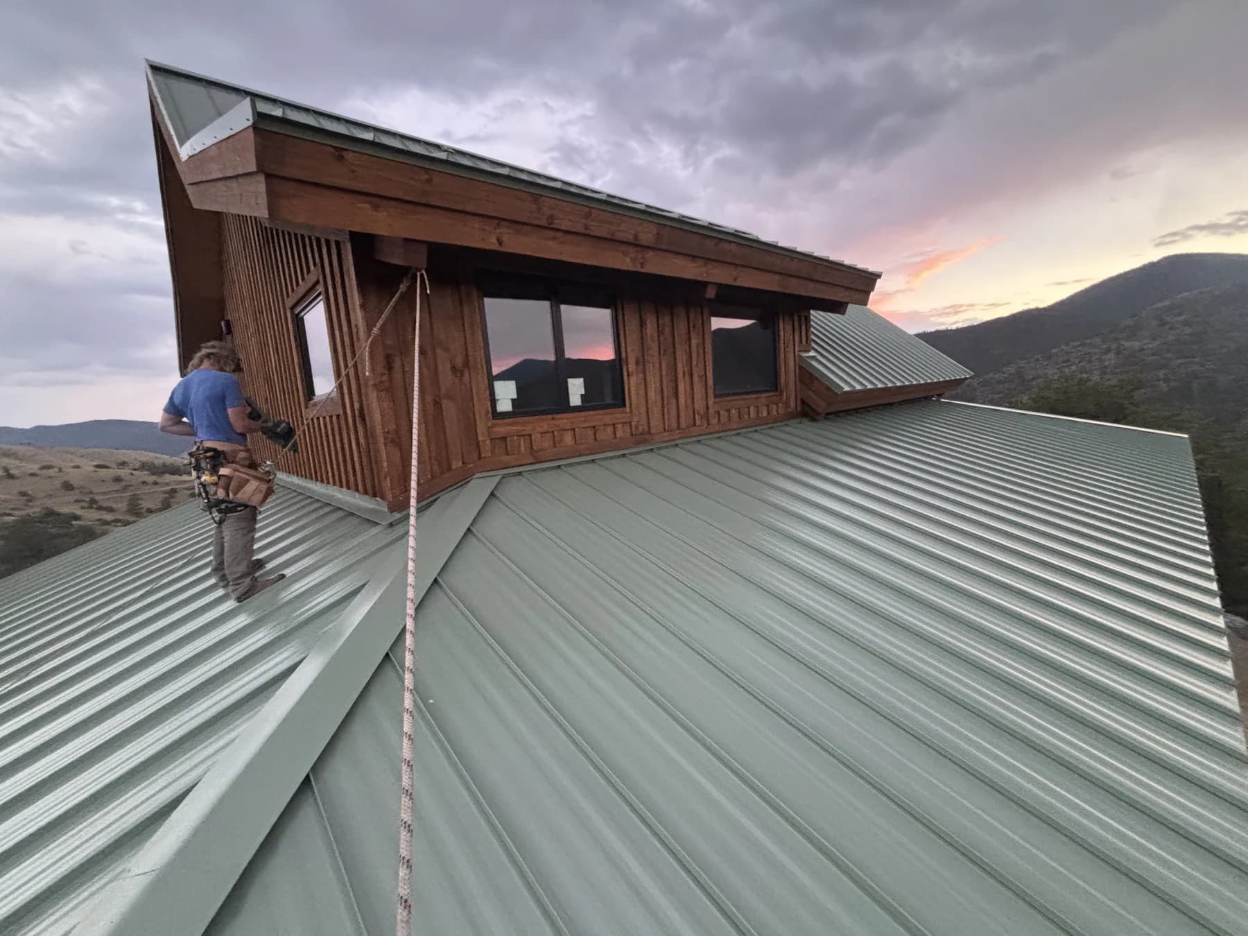 A crew member on a metal roof beside intersecting roof planes and wood siding
