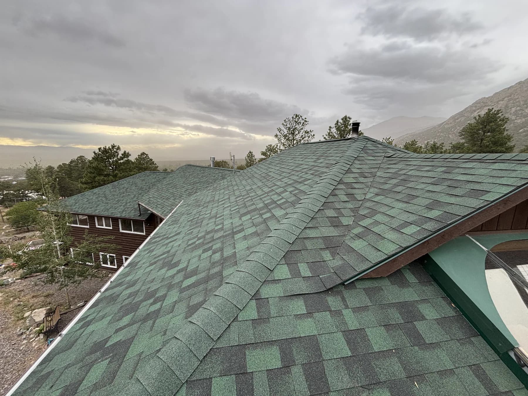 Green asphalt-shingle roofing across several roof planes in a mountain setting.
